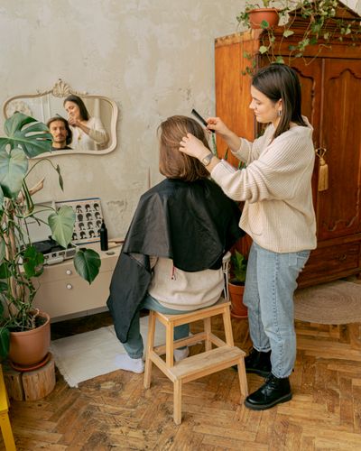 A stylist cutting a seated client's hair in a cosy, plant-filled home studio